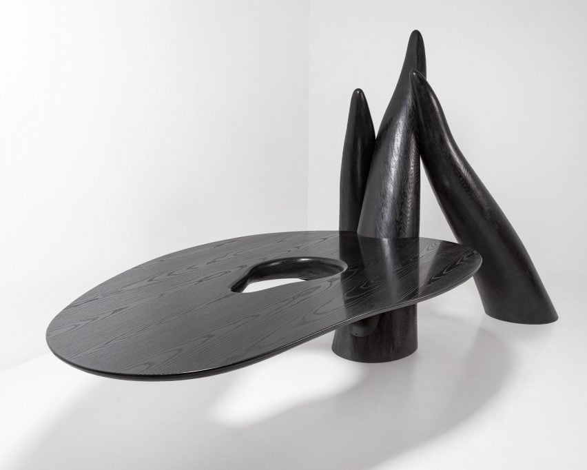 Фотография черного скульптурного объекта с большой поверхностью, напоминающей столешницу, выступающей из трех колонн, похожих на щупальца, возвышающихся над полом.